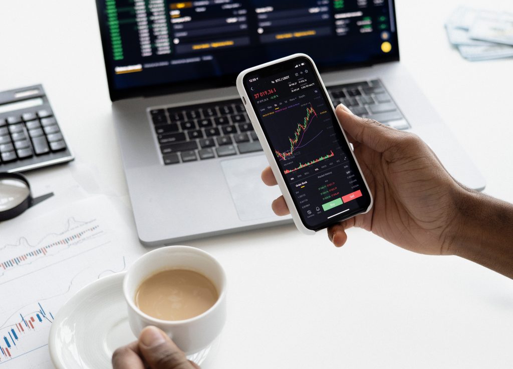 LearnOnline Self-study Economics A level Hand holding phone with finance market data with laptop in the background