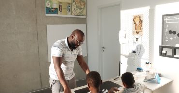LearnOnline supporting home educating families Father stood with two children sat at desk writing with educational information on the walls