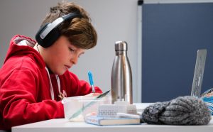 LearnOnline IGCSE Packages 14-16 year old boy with headphones on, writing in book in front of laptop